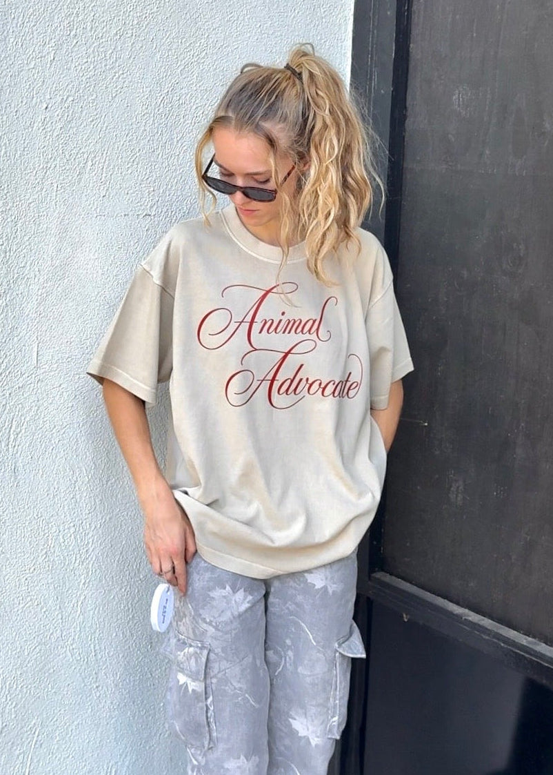 A woman wearing an oversized beige graphic t-shirt with 'Animal Advocate' printed in red cursive, paired with gray tie-dye cargo pants and white sneakers, standing next to a white textured wall.